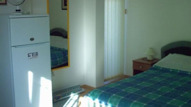 Ein Schlafzimmer mit einem Bett, einem Nachttisch, einem Spiegel, einem Fenster mit Jalousien und einem Kühlschrank.
