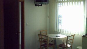 Ein Zimmer mit einem runden Tisch, vier Stühlen, einem an der Wand befestigten Fernseher und einem Fenster mit vertikalen Jalousien.