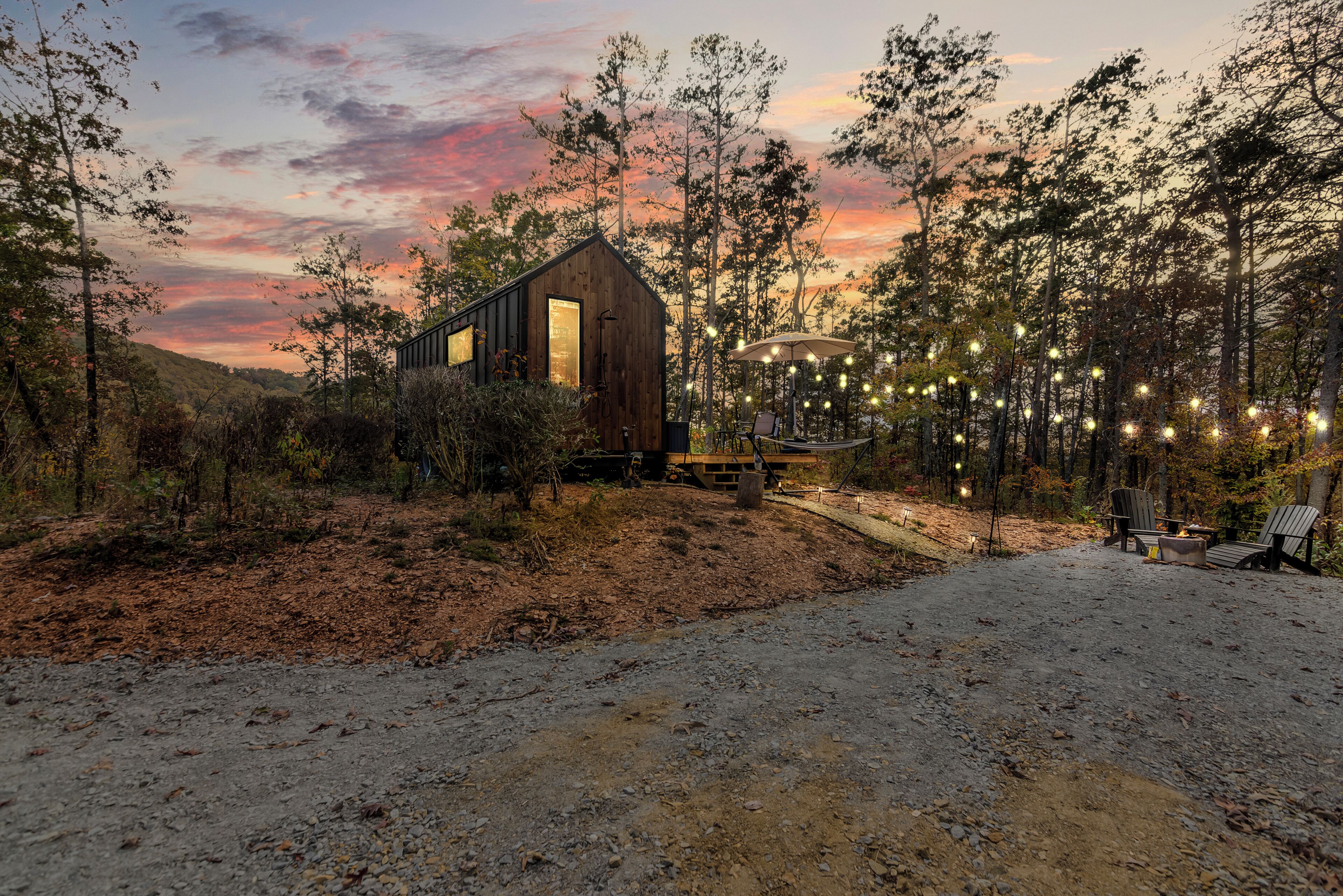 A rustic cabin with a large window, surrounded by trees and a dirt path leading to a seating area with string lights.