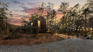 A rustic cabin with a large window, surrounded by trees and a dirt path leading to a seating area with string lights.