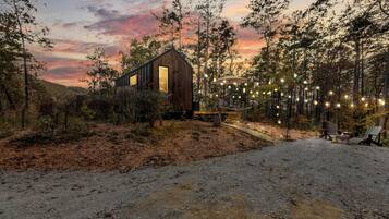 A rustic cabin with a large window, surrounded by trees and a dirt path leading to a seating area with string lights.