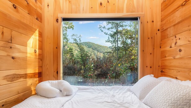A wooden cabin room with a bed, pillows, and a window overlooking a forested hillside.