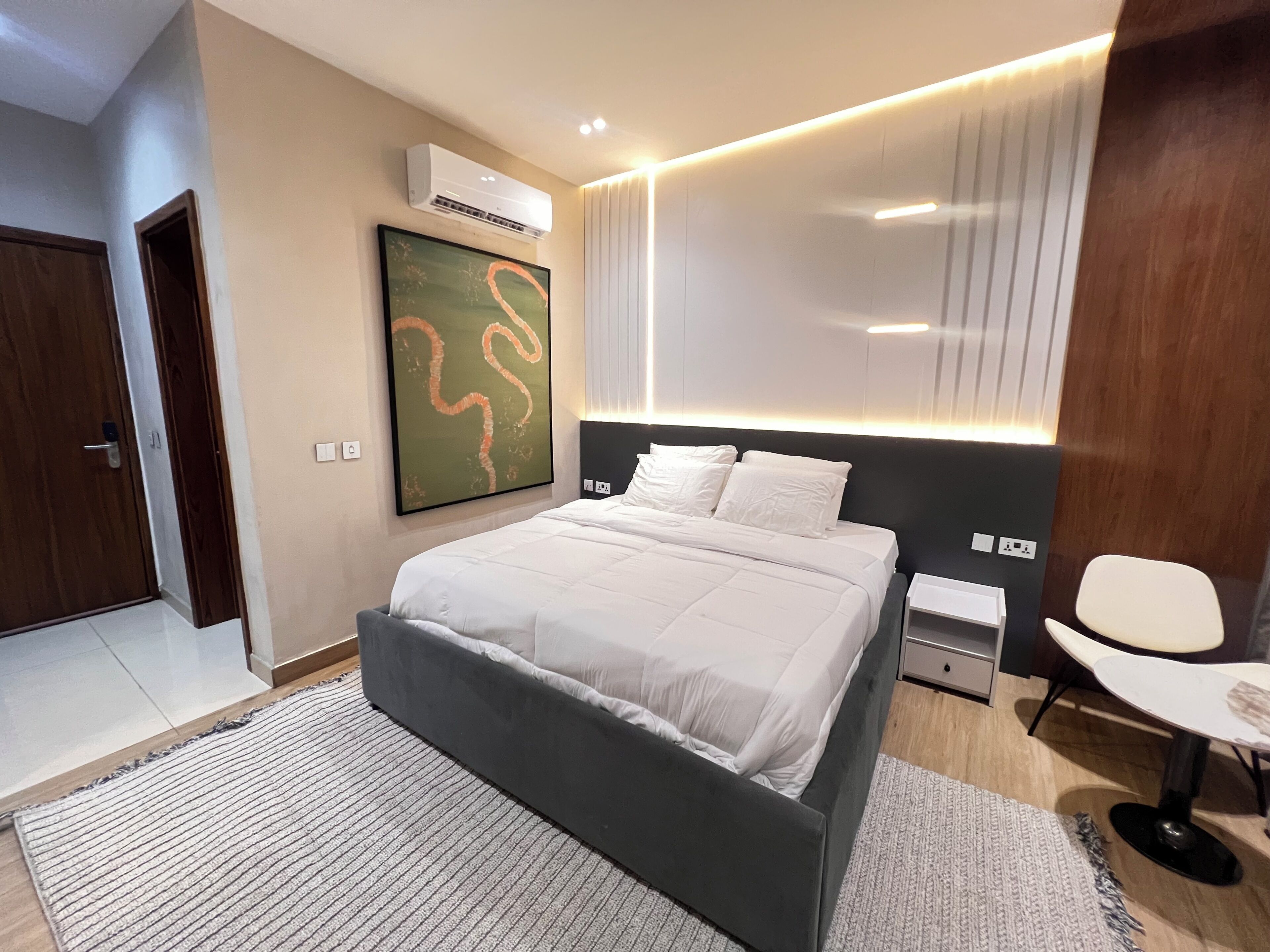 A modern bedroom with a large bed, a wall-mounted air conditioner, a painting, and a bedside table with a lamp.