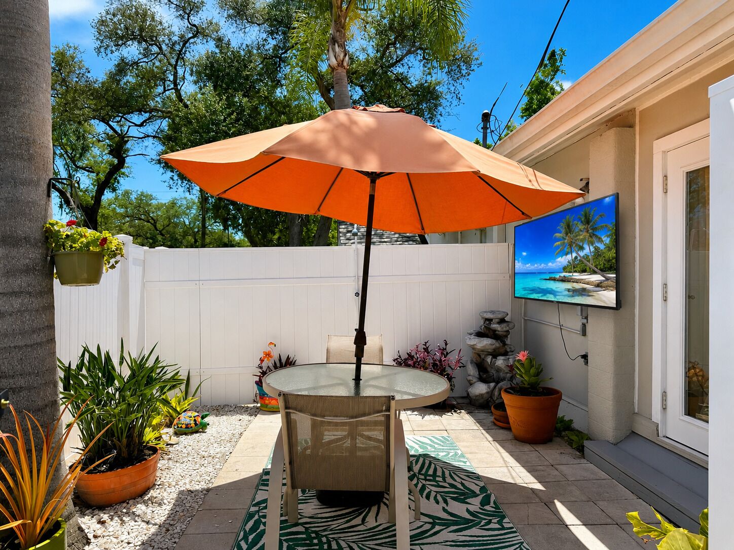 Eine Terrasse mit einem runden Tisch, einem orangefarbenen Sonnenschirm und verschiedenen Kübelpflanzen.