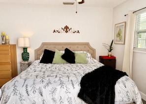 A bedroom with a tufted headboard, a bed with patterned bedding, a bedside lamp, a decorative wall piece, a window with blinds, and a potted plant.