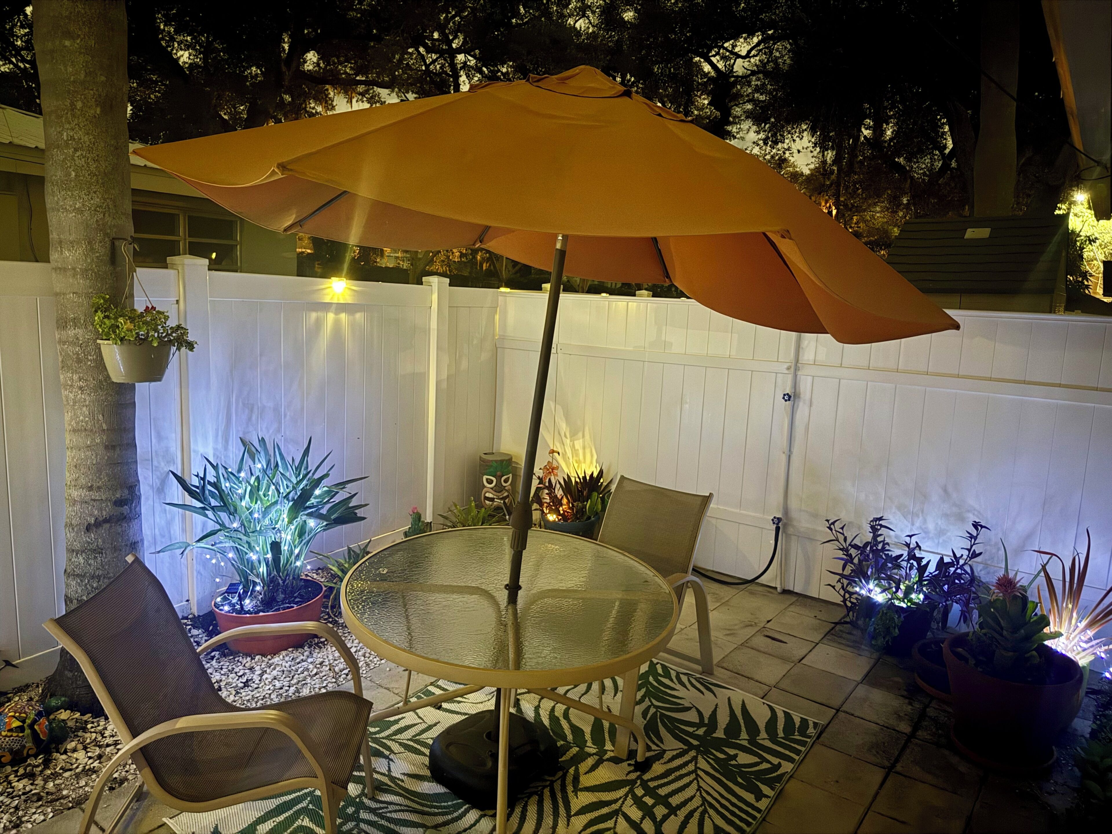 Une terrasse extérieure avec une table ronde en verre, deux chaises, un grand parasol et des plantes en pot.