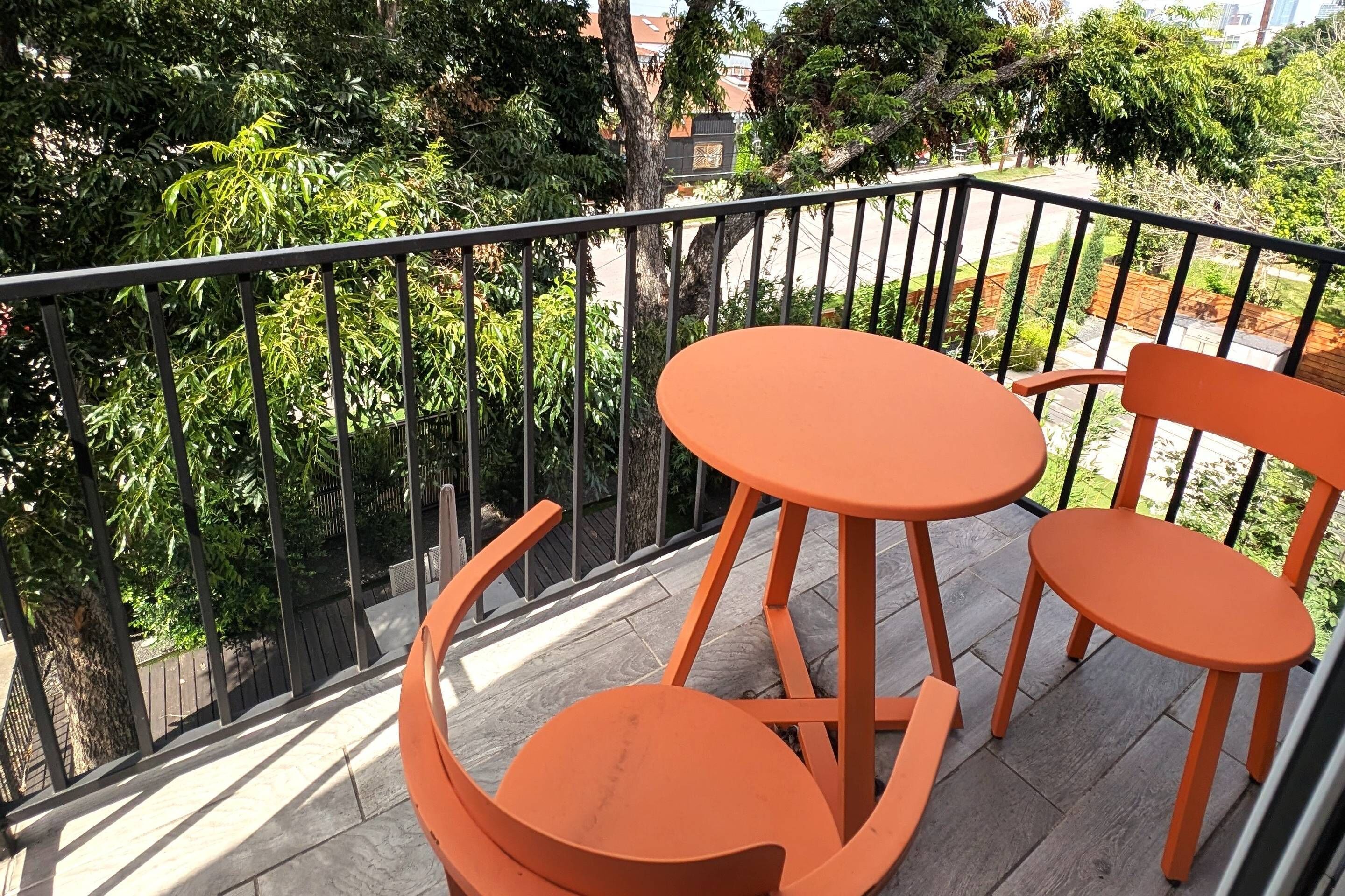 Ein Balkon mit Tisch und Stühlen, umgeben von einem Metallgeländer, mit Blick auf eine Wohngegend mit Bäumen und Häusern.