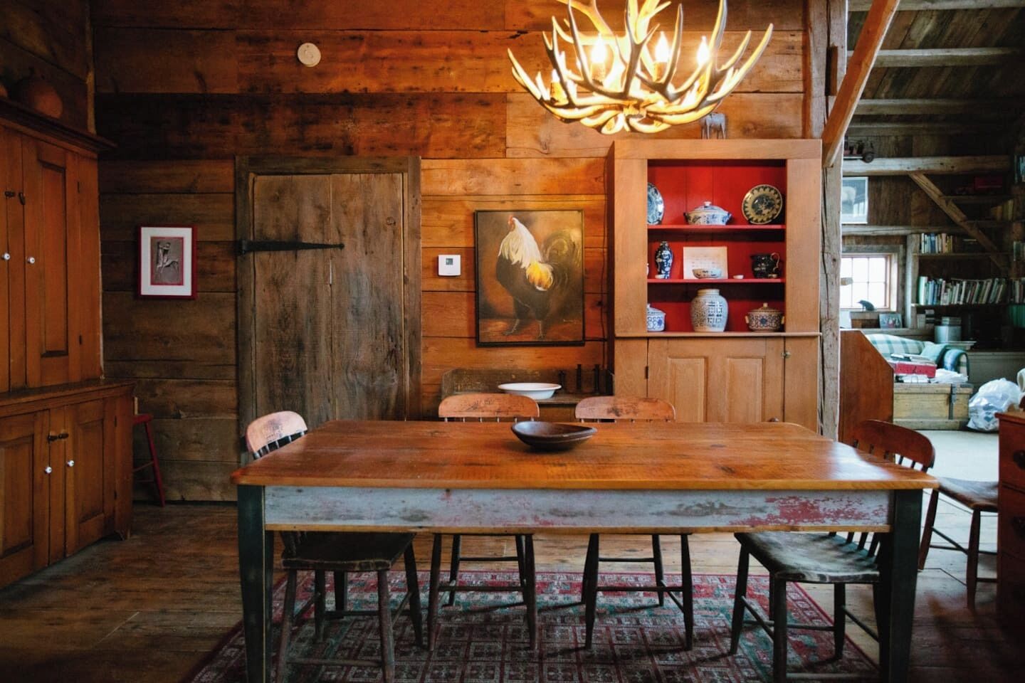 A rustic wooden dining area with a large table, chairs, a chandelier, and a bookshelf with decorative items.