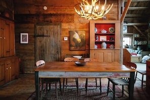 Un coin repas rustique en bois, avec une grande table, des chaises, un lustre et une bibliothèque agrémentée d’objets décoratifs.