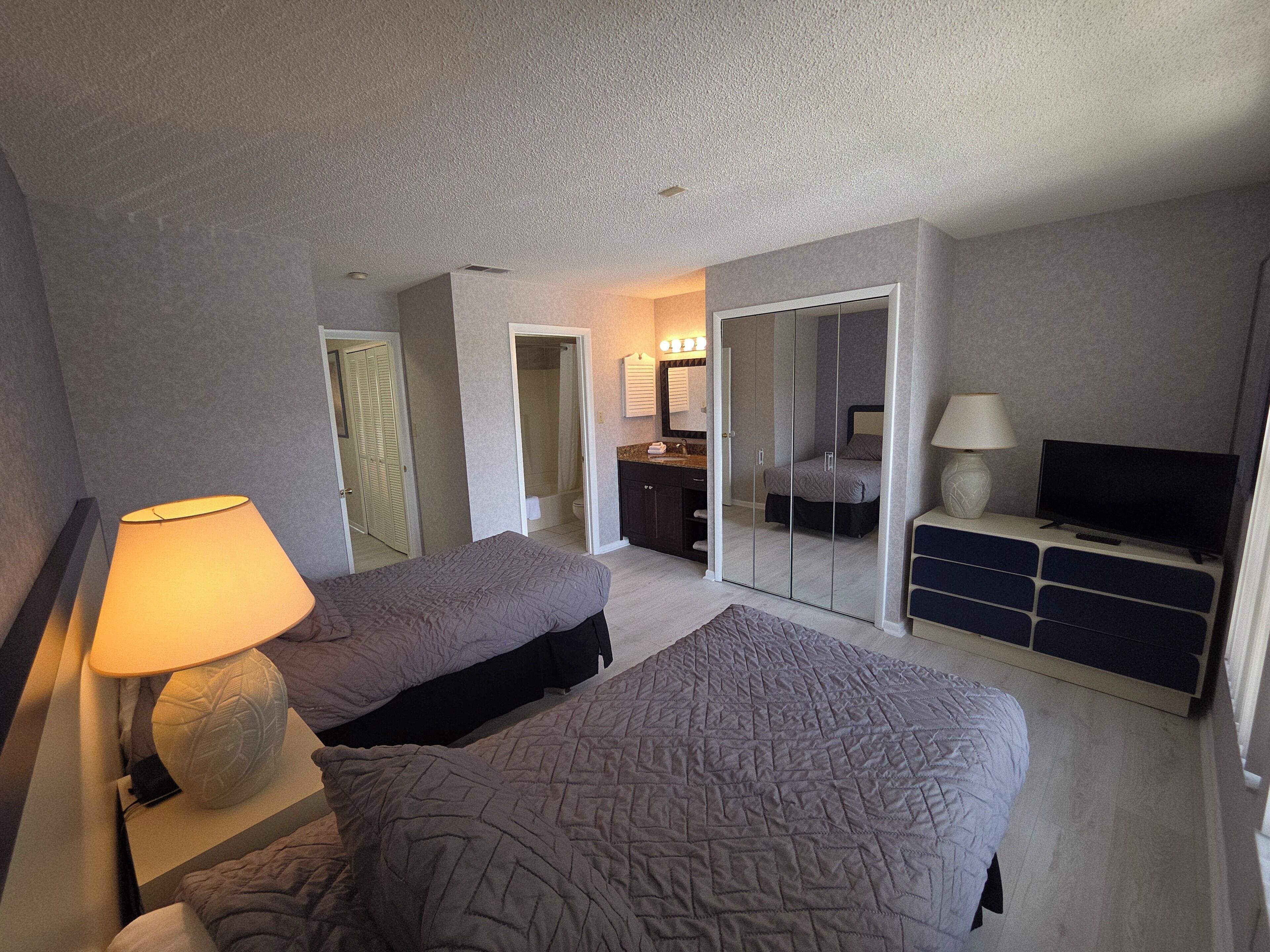 Habitación de hotel con dos camas, televisor, escritorio y baño visible a través de una puerta abierta.