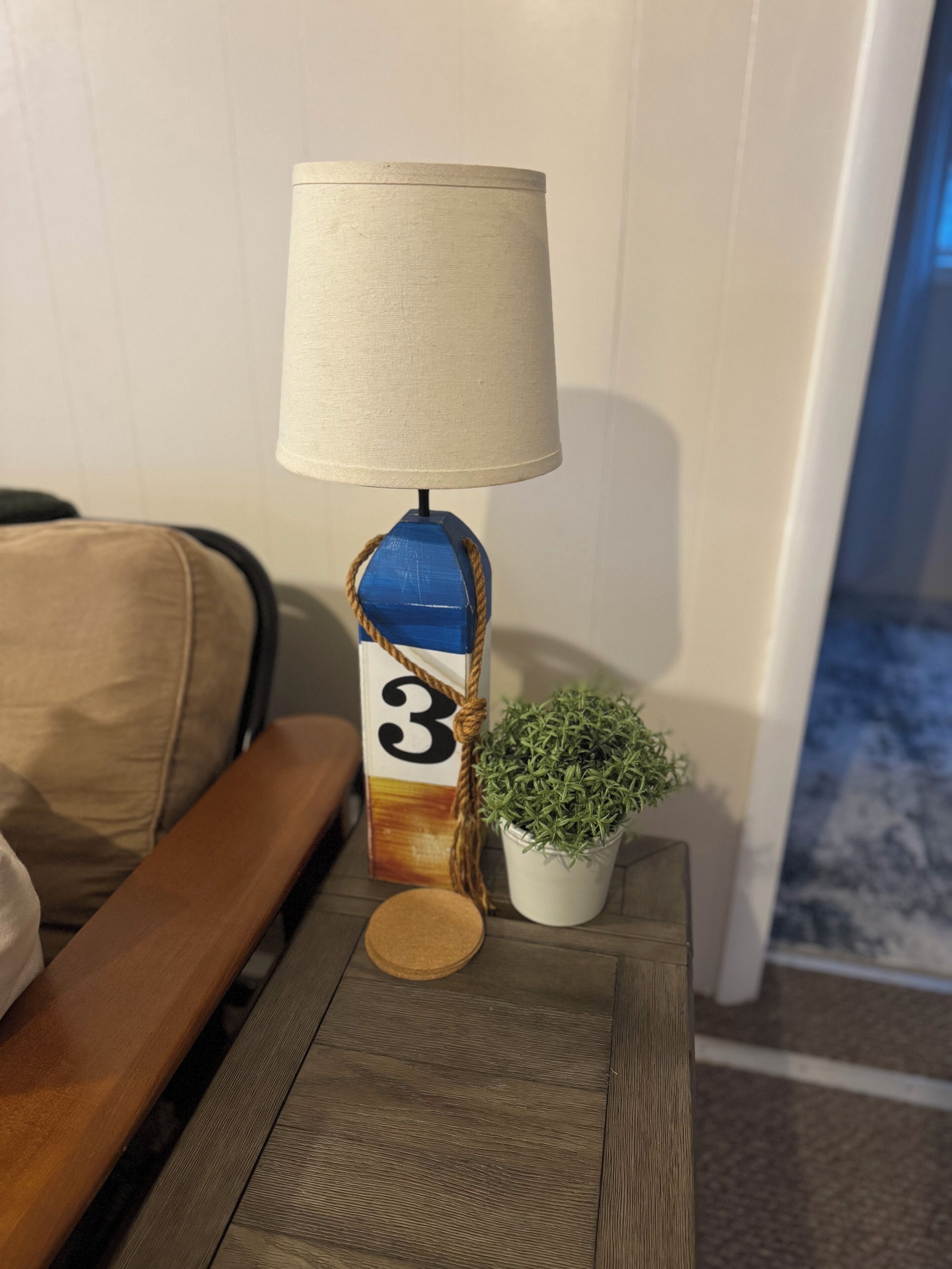 A lamp with a beige shade, a blue base with a number 3, and a small potted plant on a wooden table.