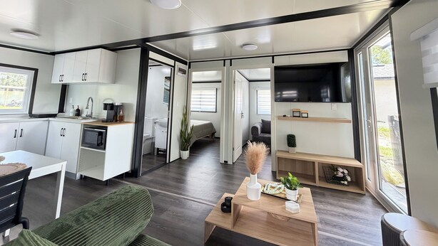Ein modernes Interieur mit Küche, Essbereich und Wohnbereich, ausgestattet mit einem großen Fernseher, Holzböden und einer Schiebetür, die zu einem Schlafzimmer führt.