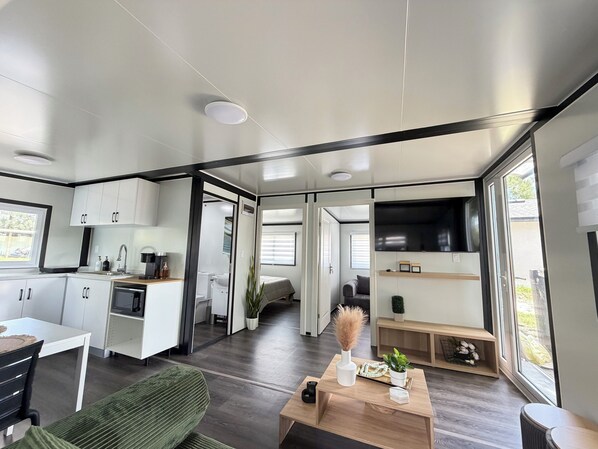 Un interior moderno con cocina, comedor y sala de estar, que incluye un televisor grande, pisos de madera y una puerta corredera que da a un dormitorio.