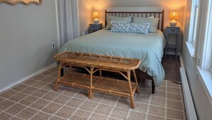 Une chambre à coucher comprenant un lit, deux tables de chevet, un miroir, une fenêtre et un banc en rotin.