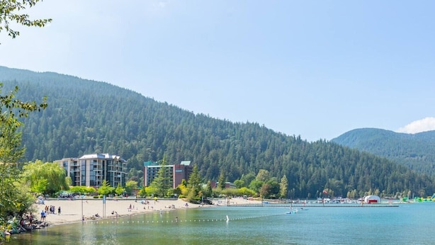 Hotel aan een meer met een strand, beboste heuvels op de achtergrond en een heldere lucht.