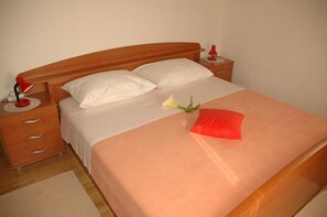Ein ordentlich bezogenes Bett mit einem Holz-Kopfteil, weißen Kissen, einer pfirsichfarbenen Tagesdecke und einem roten Kissen, sowie einer einzelnen weißen Blume.