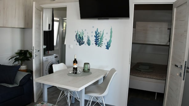 A compact living space with a dining table, chairs, a sofa, a television mounted on the wall, and a washing machine in the background. - Mobile Home With Terrace And Sea View (Pakostane)