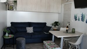 A compact living space with a dark blue sofa, a small round table with a vase, a white table with a bottle and glasses, and a wall-mounted TV. - Mobile Home With Terrace And Sea View (Pakostane)