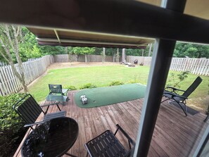 Un patio trasero con una terraza de madera, césped artificial verde y mobiliario exterior, incluyendo sillas y una mesa.