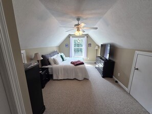 Un dormitorio con cama, cómoda, televisor, ventilador de techo y ventana.