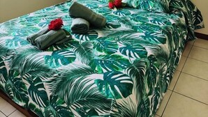 Una cama con una colcha de estampado tropical, un cuadro de una pareja en la playa y una pequeña estantería con un florero.