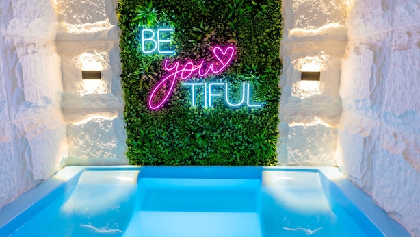Un espace de bien-être avec une piscine bleue, un néon affichant le mot « BE your TIFUL » et un mur végétal.