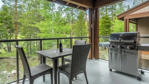 Ett takförsett utomhusområde med ett bord och stolar, en grill och utsikt över en skog.