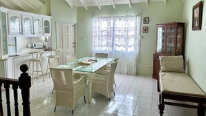 Comedor con mesa de vidrio y sillas de mimbre, cocina con muebles blancos y un armario de madera con cristalería.