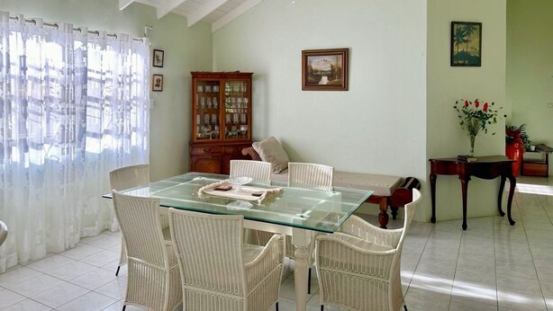 Un comedor con una mesa de vidrio, sillas de mimbre, ventilador de techo, un mueble con puertas de cristal y un cuadro en la pared.