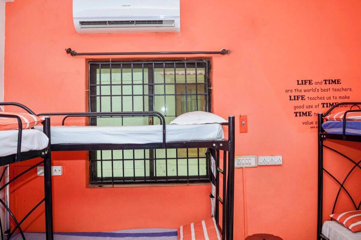 A dormitory room with bunk beds, an air conditioning unit, a barred window, and a motivational quote on the wall.