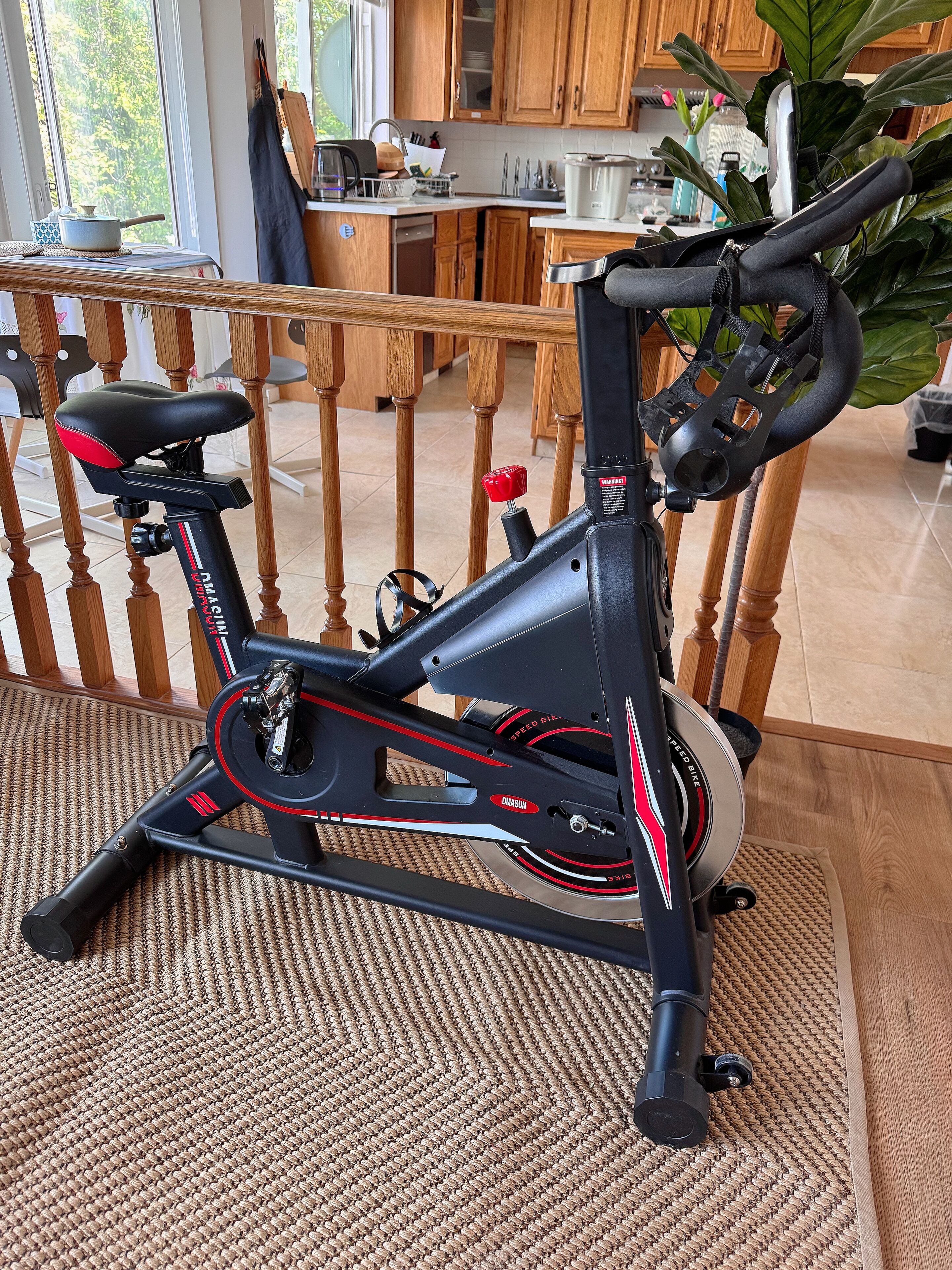 Een stilstaande hometrainer in een ruimte met houten vloeren, een keuken op de achtergrond en een raam met uitzicht op bomen.