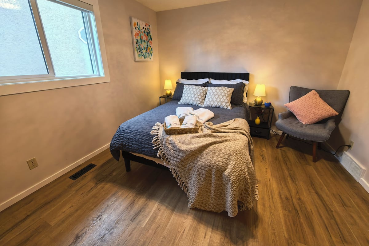 Un dormitorio con cama, dos mesitas de noche con lámparas, una silla, una ventana y un cuadro en la pared.