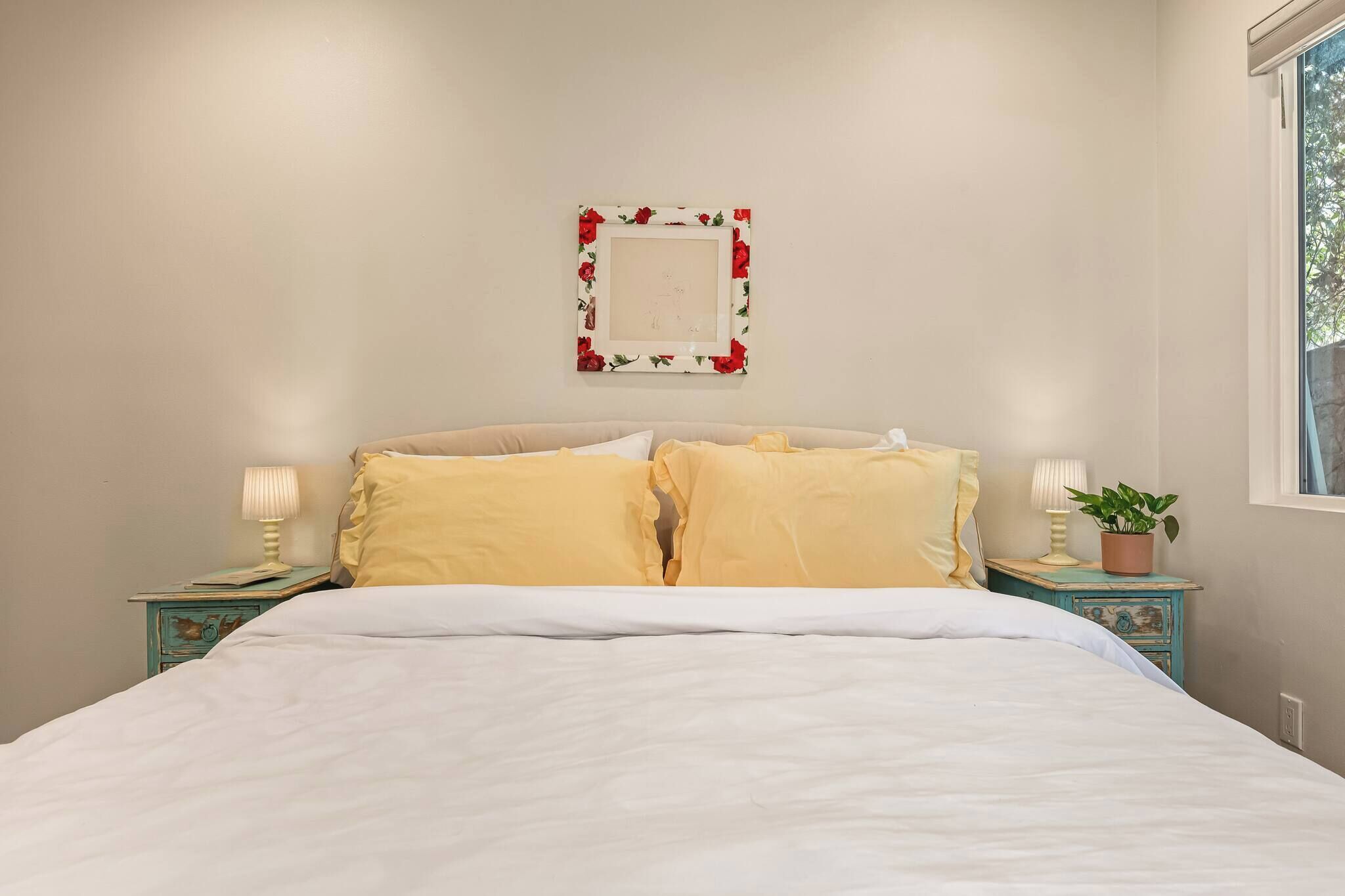 Un lit avec une literie blanche, deux oreillers jaunes, un cadre photo à motifs floraux accroché au mur, et deux tables de chevet avec des lampes et une plante.