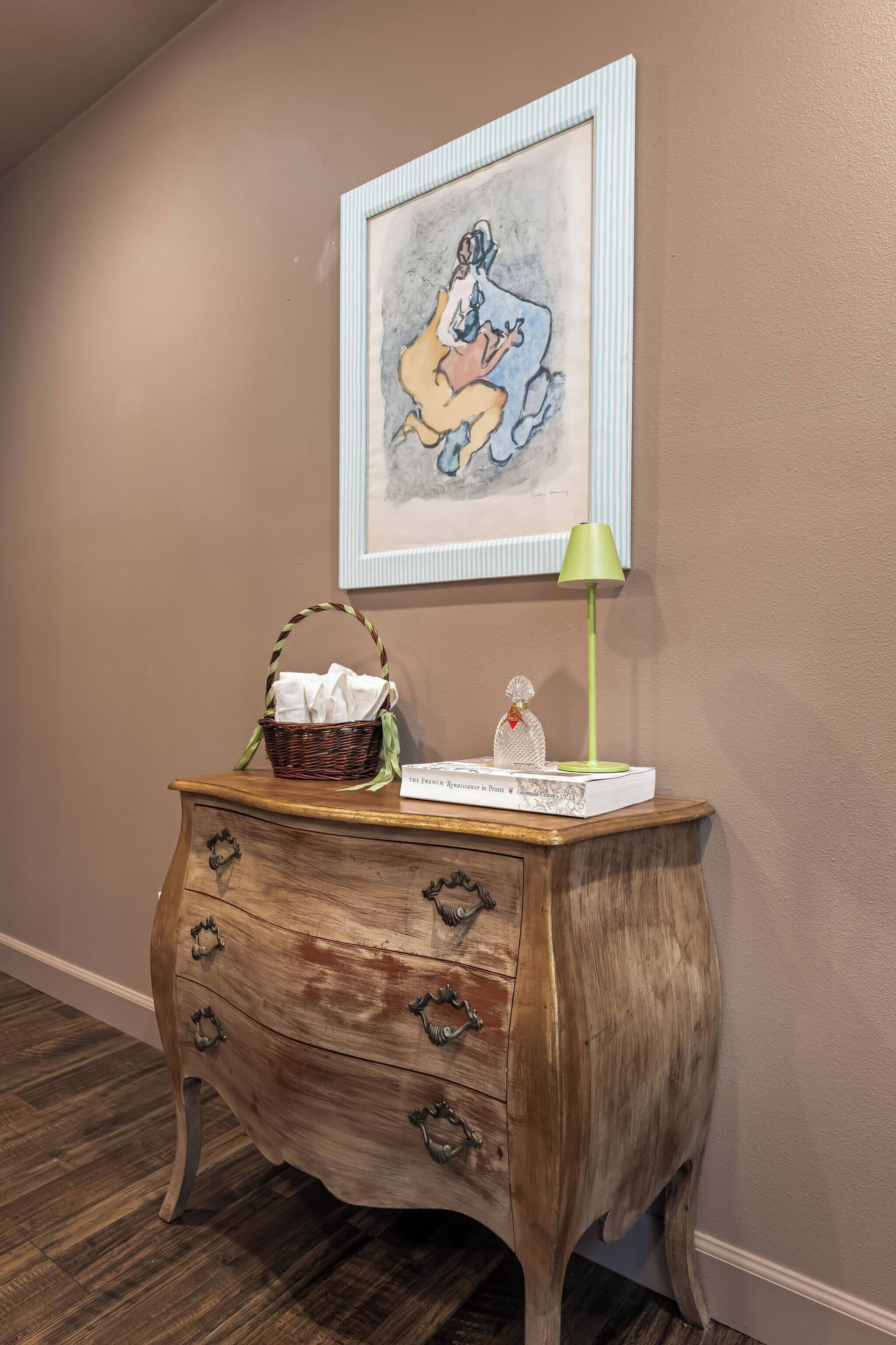 A wooden dresser with drawers, a framed picture on the wall, a basket, a green lamp, and a decorative item on the dresser.