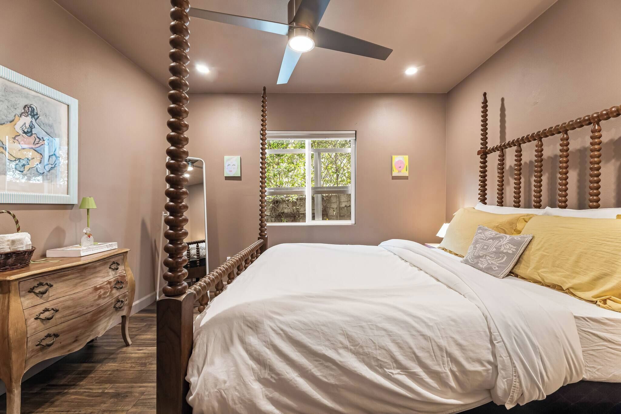 A bedroom with a four-poster bed, a ceiling fan, a window with a view of greenery, and a wooden dresser with drawers.
