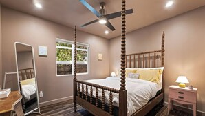 A four-poster bed with a ceiling fan, a bedside table with a lamp, a mirror, and a window with a view of trees.