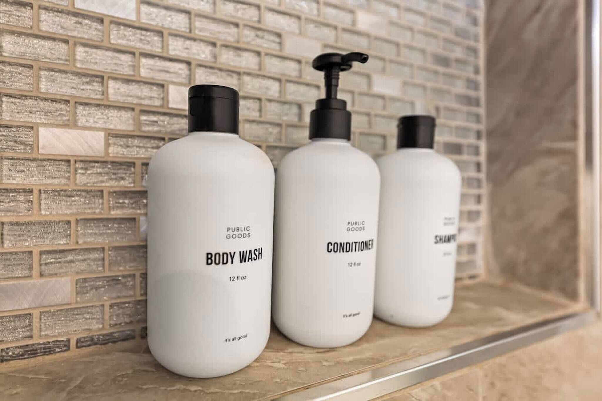 Tre hvide flasker med bodywash, balsam og shampoo står på en hylde foran en murstensvæg.