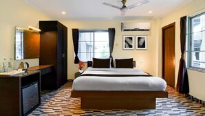 A hotel room with a bed, a desk with a coffee maker, a small refrigerator, an air conditioning unit, a ceiling fan, and a window with curtains.