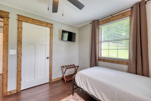 A bedroom with a bed, a ceiling fan, a TV, a window with blinds, and a door.