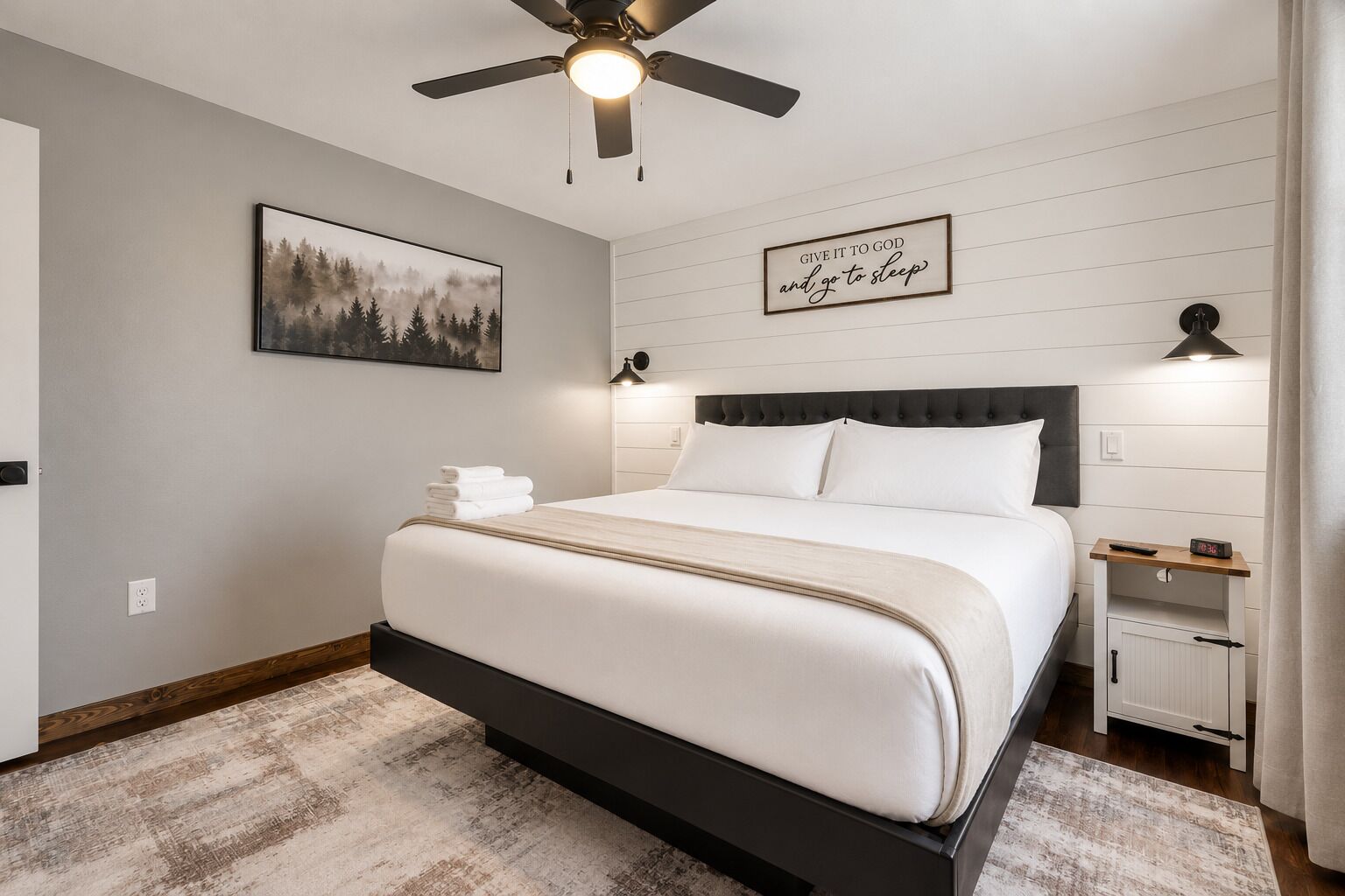A bedroom with a bed, a ceiling fan, a bedside table with a clock, and a wall-mounted light fixture.