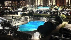 Ein Schwimmbecken mit Liegestühlen, einem überdachten Bereich und Blick auf einen Parkplatz sowie Gebäude im Hintergrund.