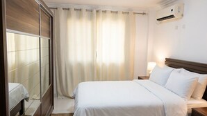 Hotellihuone, jossa on sänky, yöpöytä, seinään asennettu ilmastointilaite ja ikkuna, jossa on verhot.
