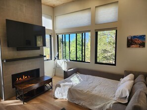 Ein modernes Schlafzimmer mit Kamin, einem Bett, einem Fernseher und großen Fenstern mit Blick auf Bäume.