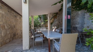 An outdoor patio area with stone flooring, a long table, wicker chairs, and a view of a rocky cliff. - Amazing Villa in Omis With Private Pool (Omis)