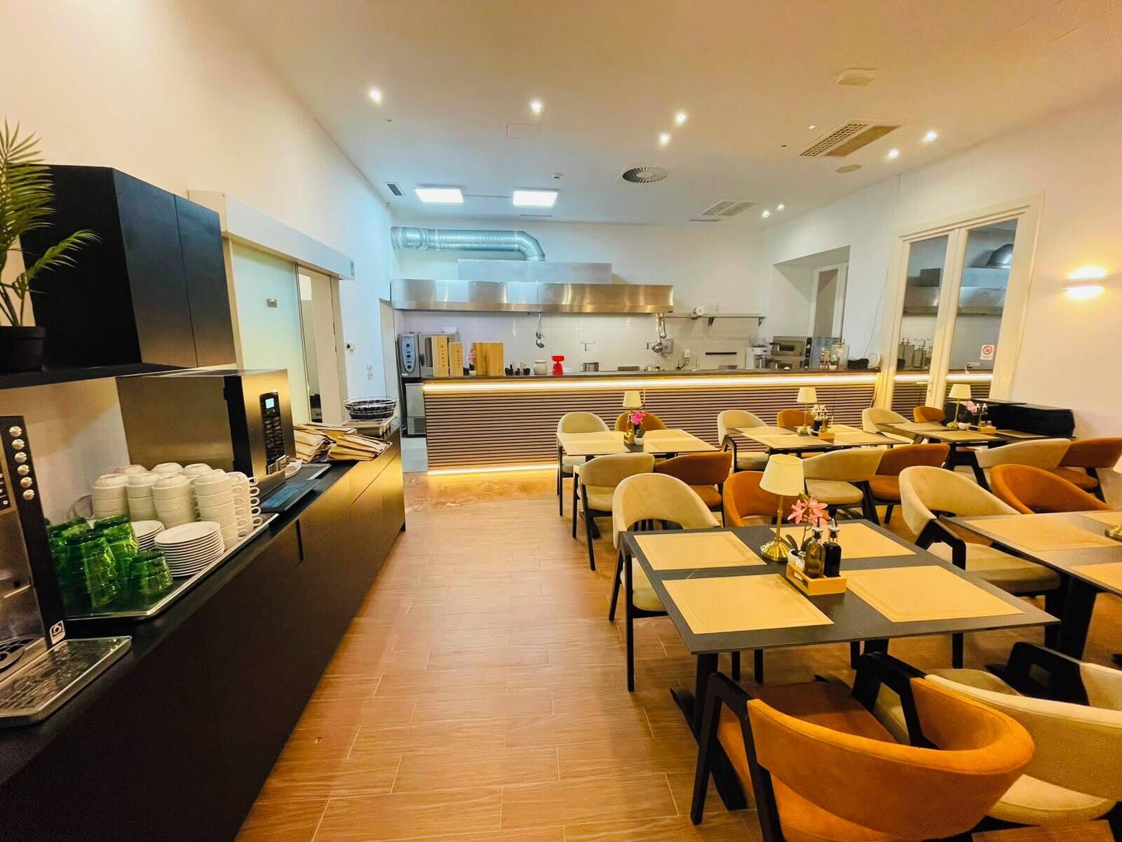 Moderni ruokailutila, jossa on pöytiä ja tuoleja, taustalla keittiö ja vasemmalla puolella buffetpöytä.