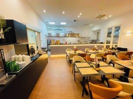 Un comedor moderno con mesas y sillas, una cocina al fondo y un mostrador de buffet a la izquierda.