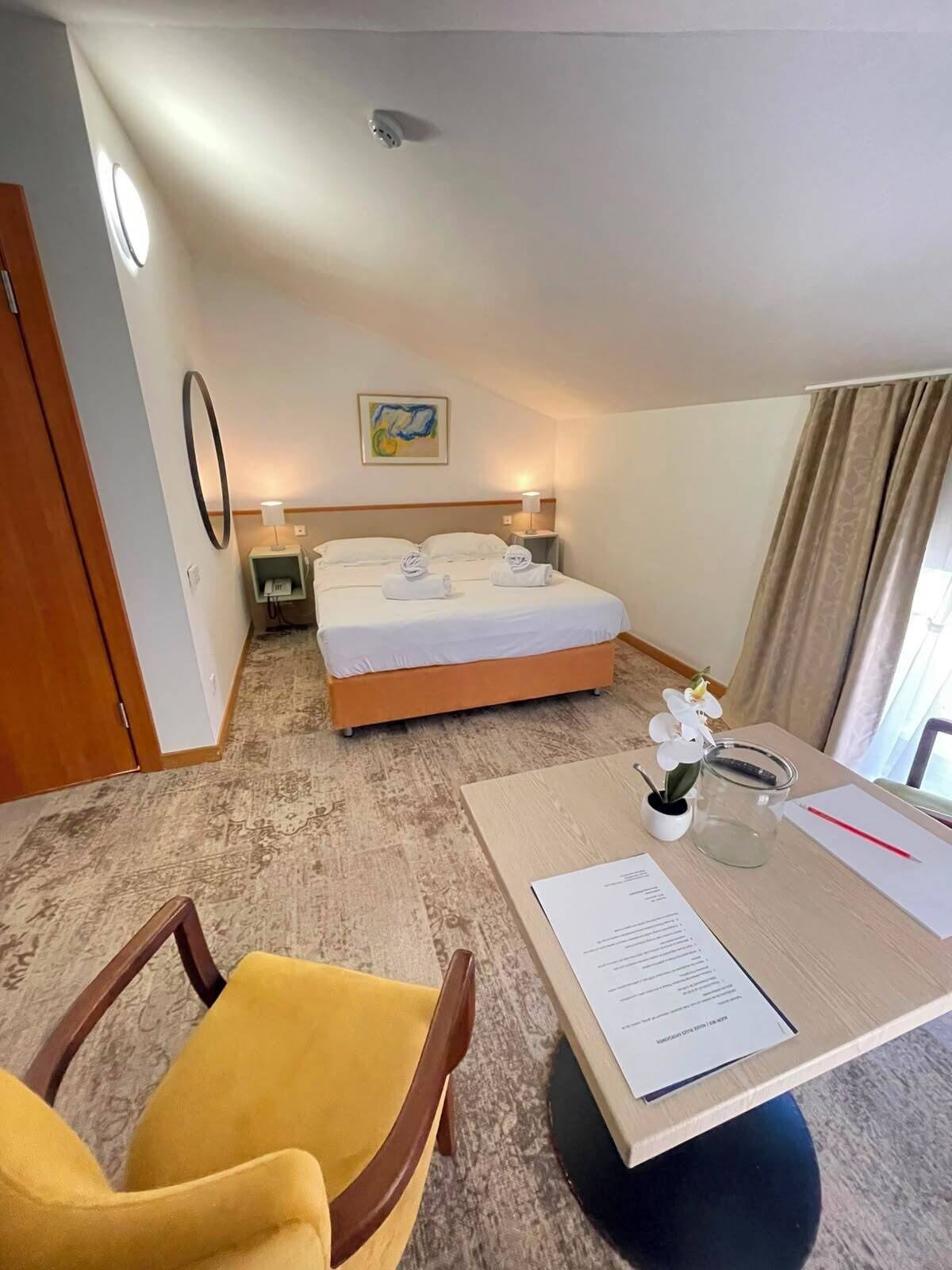 Une chambre d’hôtel comprenant un lit, un bureau avec une chaise, une petite table avec un vase et une plante, et un téléphone fixé au mur.