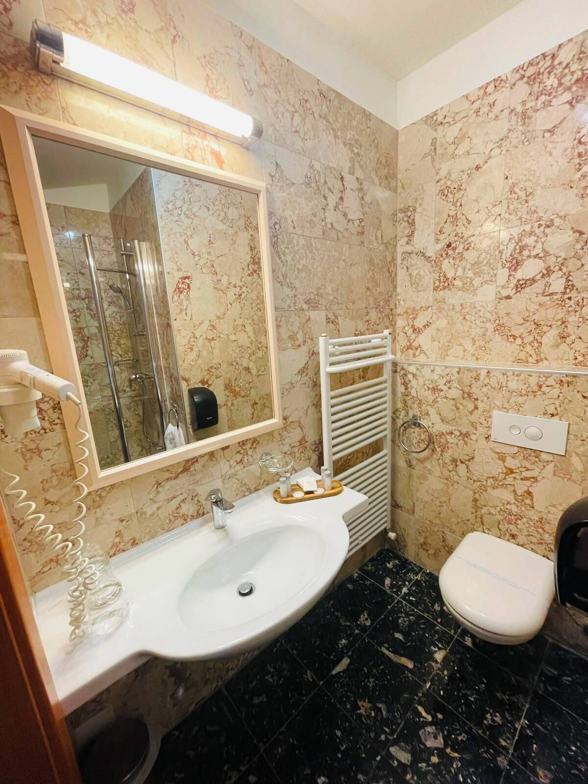 Ein Badezimmer mit Marmorwand, Waschbecken in Weiß, Spiegel, Handtuchhalter und Toilette.