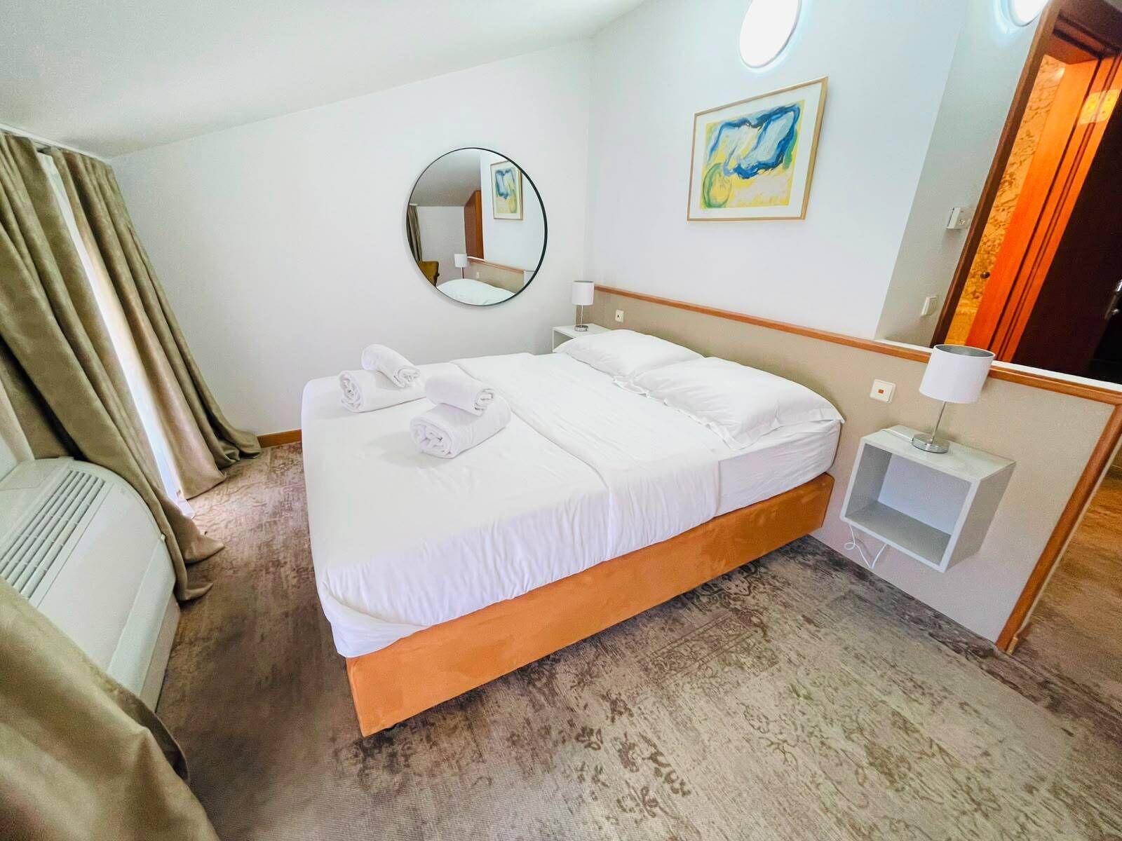 A hotel room with a bed, a bedside table with a lamp, a mirror, and a painting on the wall.