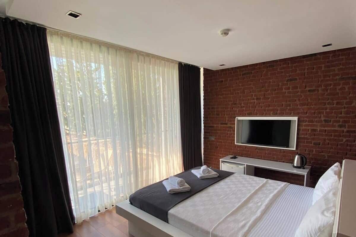 Un dormitorio moderno con una cama grande, un televisor montado en una pared de ladrillo y un balcón con cortinas.